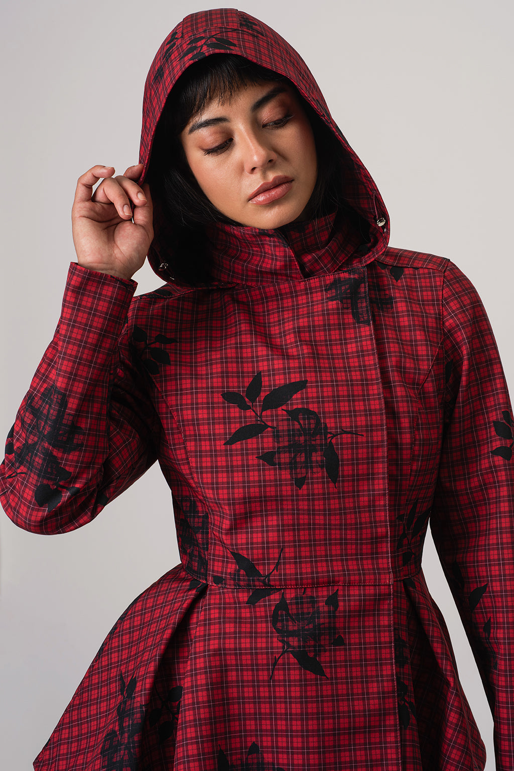 A model wearing RainSisters red and black plaid hooded coat Crimson Heart with black floral motifs, posed against a neutral light gray background.