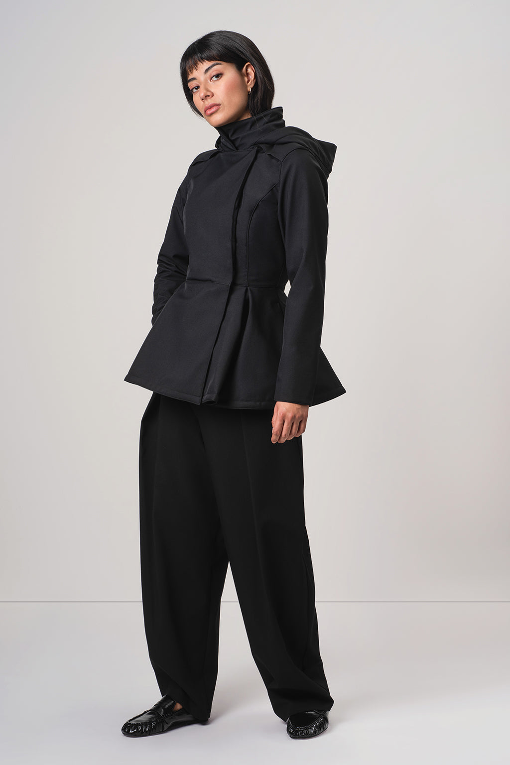 A model wearing a black RainSisters peplum-style jacket Moonlit Bow with detachable hood and scarf and matching wide-leg trousers, photographed against a neutral light gray studio background.