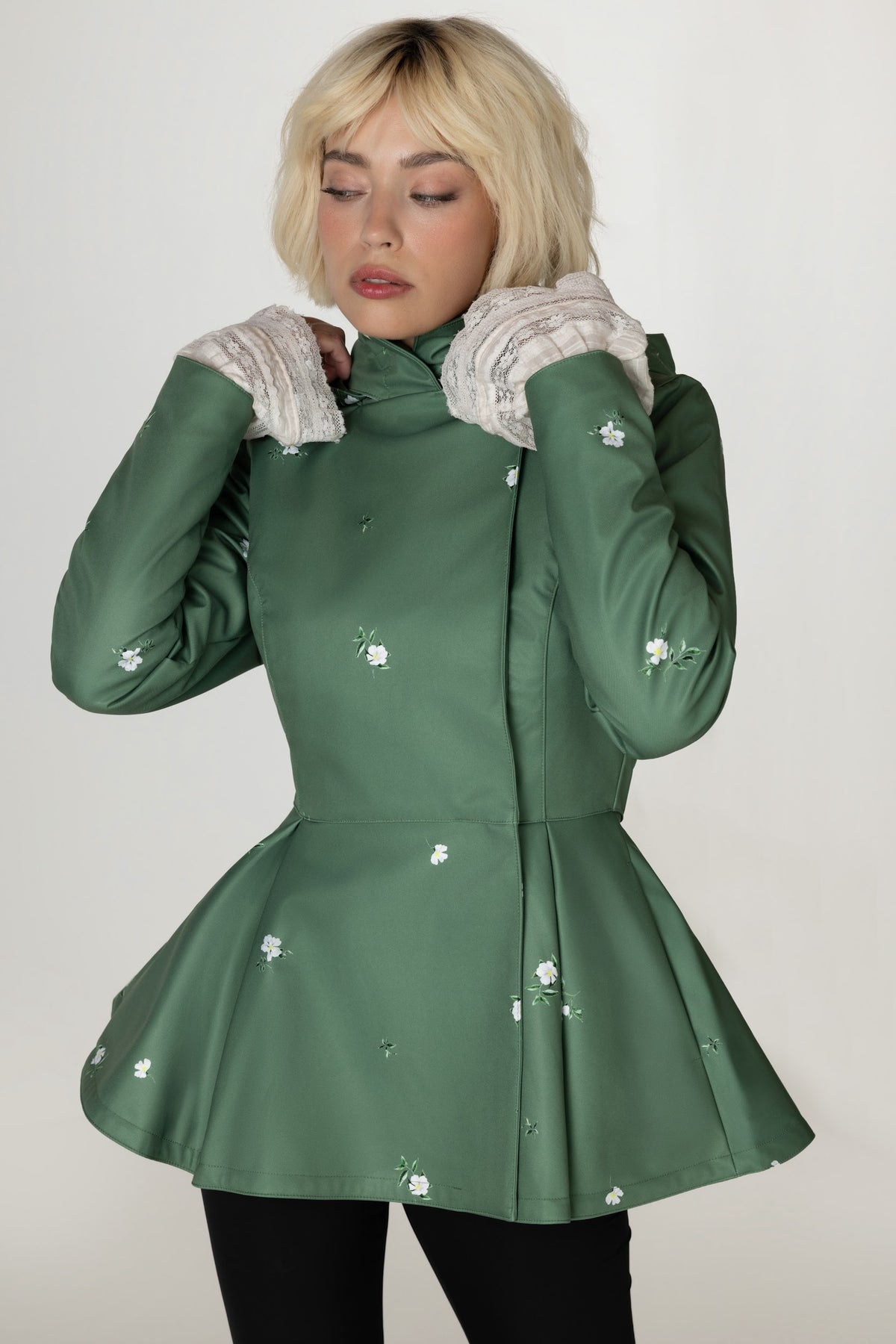 A woman wearing RainSisters green peplum-style coat with white floral print and white knit cuffs, posed against a plain light gray studio background.