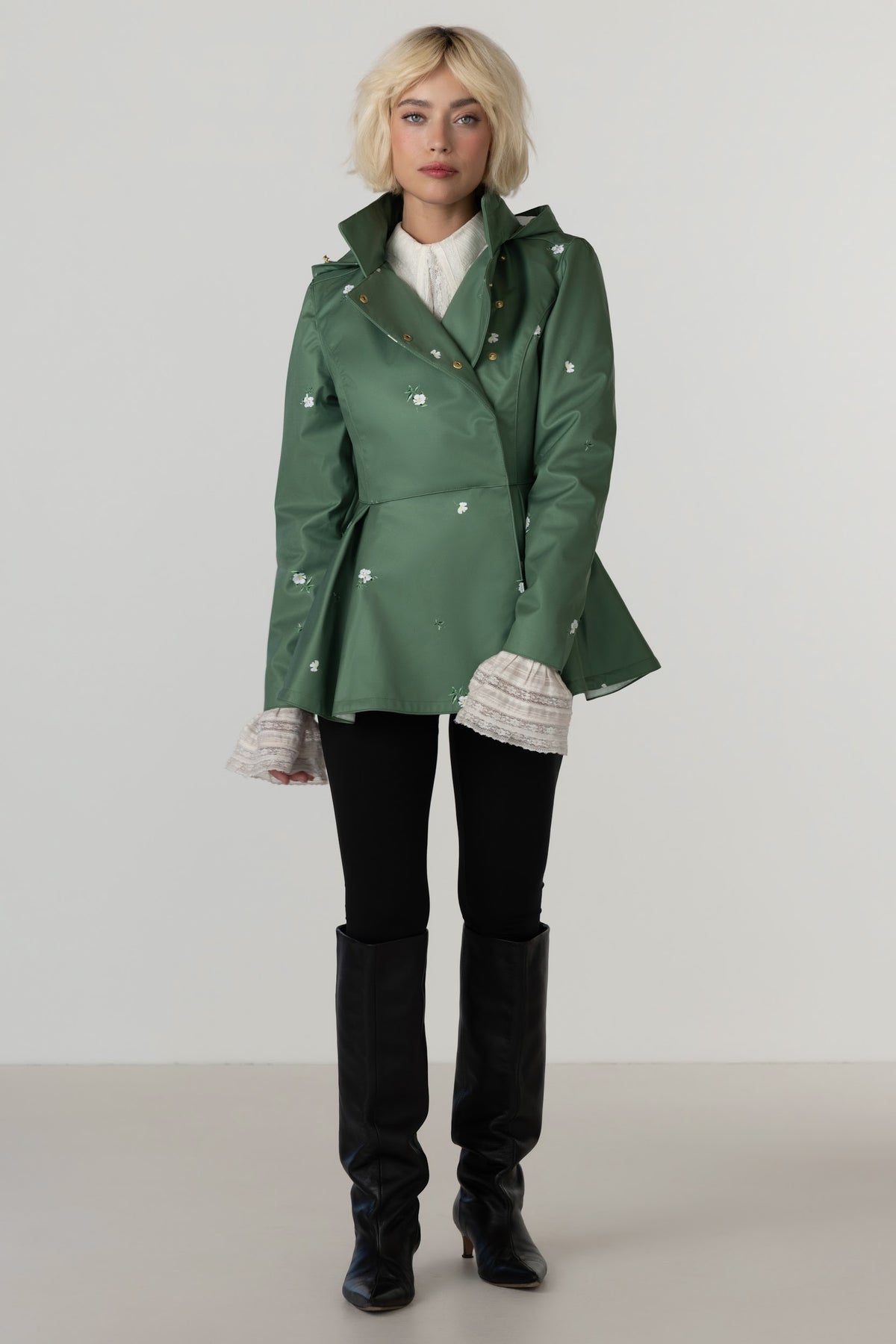 A model wearing RainSisters green double-breasted peplum-style coat Primrose Green with white floral print, styled with a white top, black pants, and black knee-high boots against a neutral studio background.