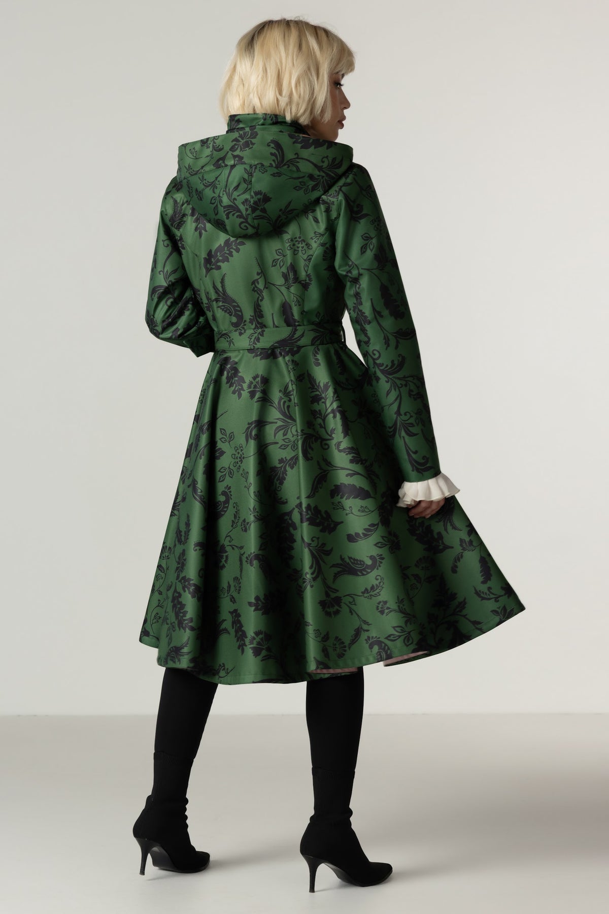 A woman shown from the back wearing a green hooded winter coat with a dark floral print, black tights, and black high heels against a light gray studio background.