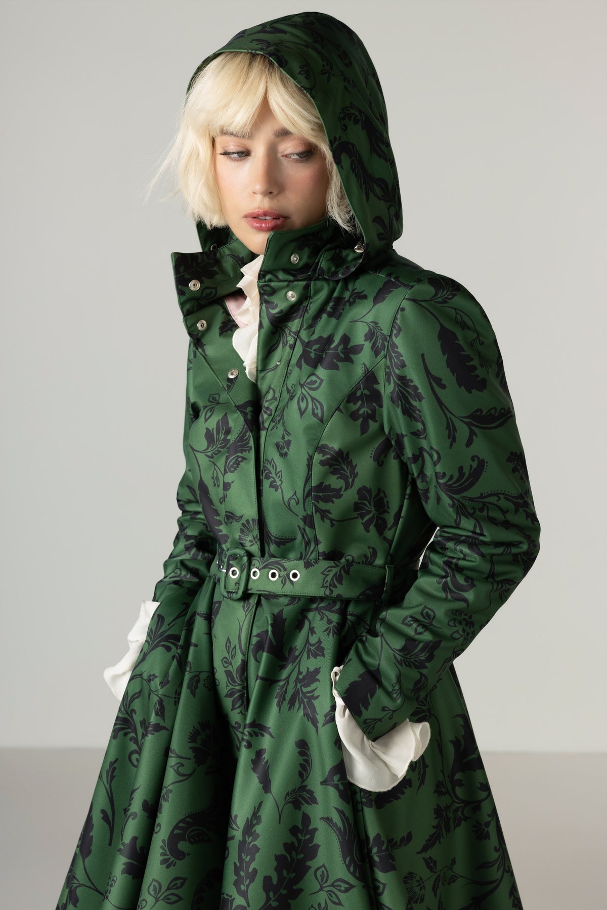 A model wearing a dark green hooded winter raincoat with a black floral pattern, featuring a matching belt and ash pink inner lining, set against a plain light gray background.