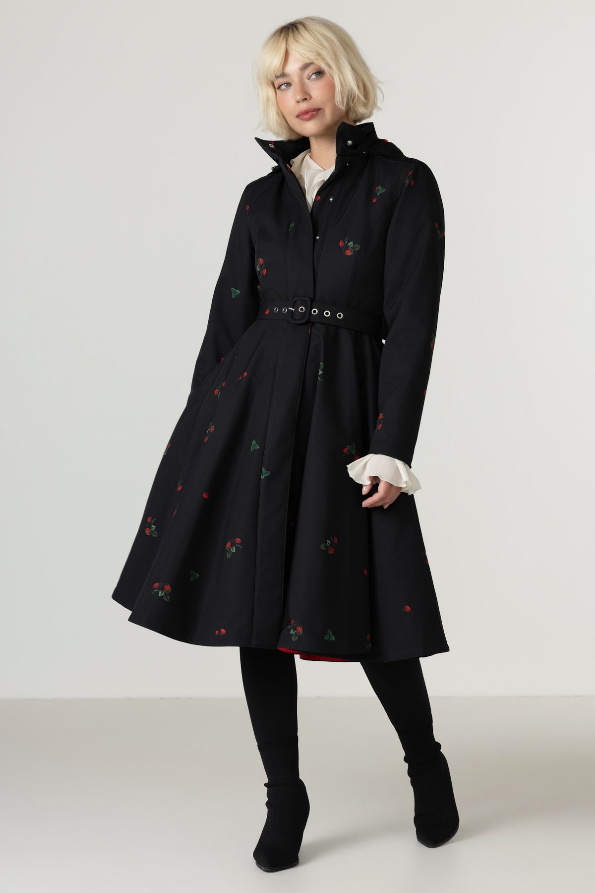 A model wearing a black knee-length coat with a high collar, a matching belt, and small red and green berry details, styled with a white shirt, black tights, and black ankle boots against a light gray studio background.