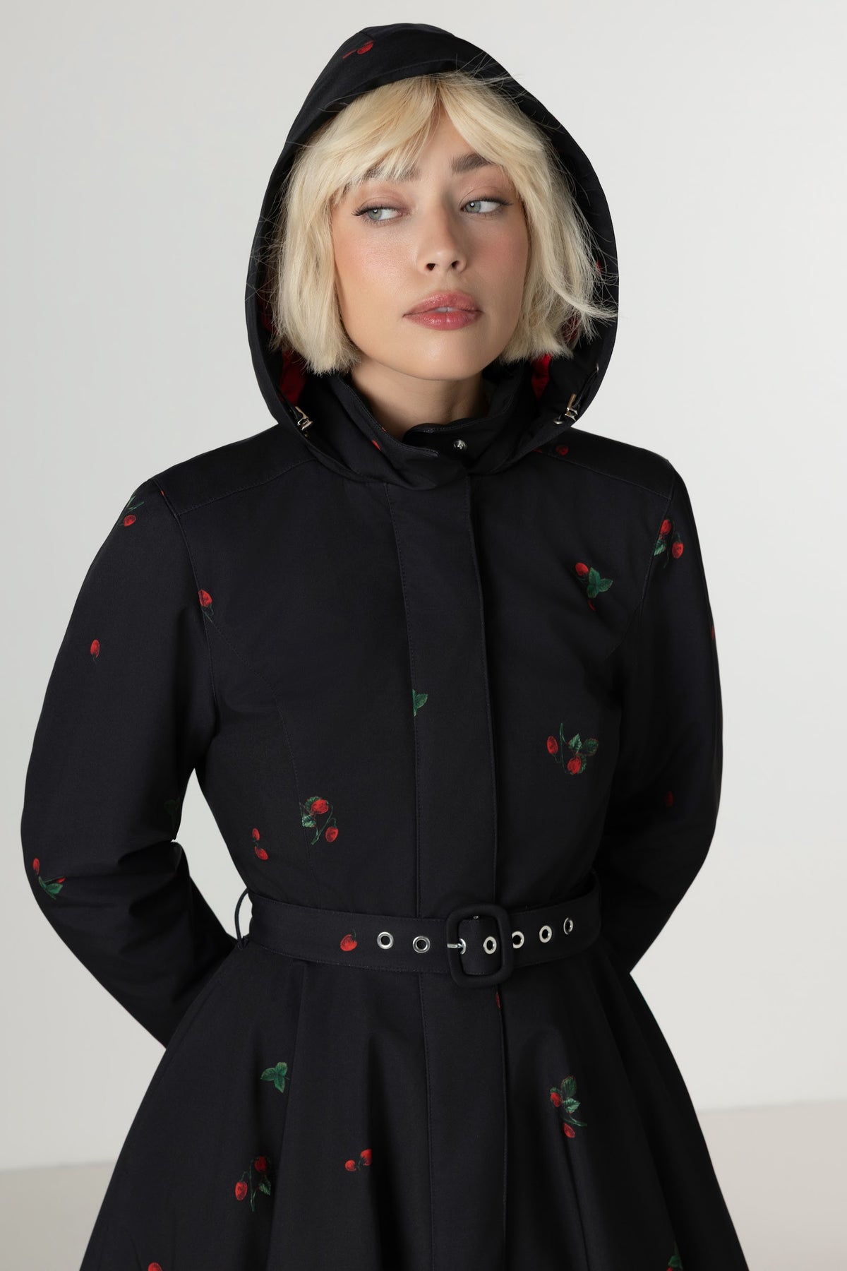 A model wearing a black hooded coat with a matching belt, decorated with small red and green berry motifs, set against a plain light gray background.