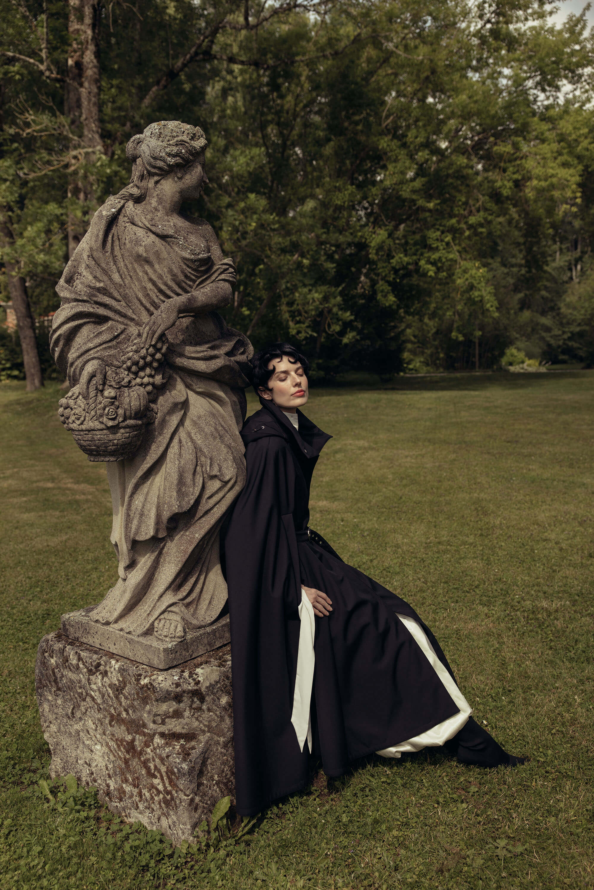 A person in a long black cloak by RainSisters reclines beside a stone statue on a grassy lawn with trees in the background.