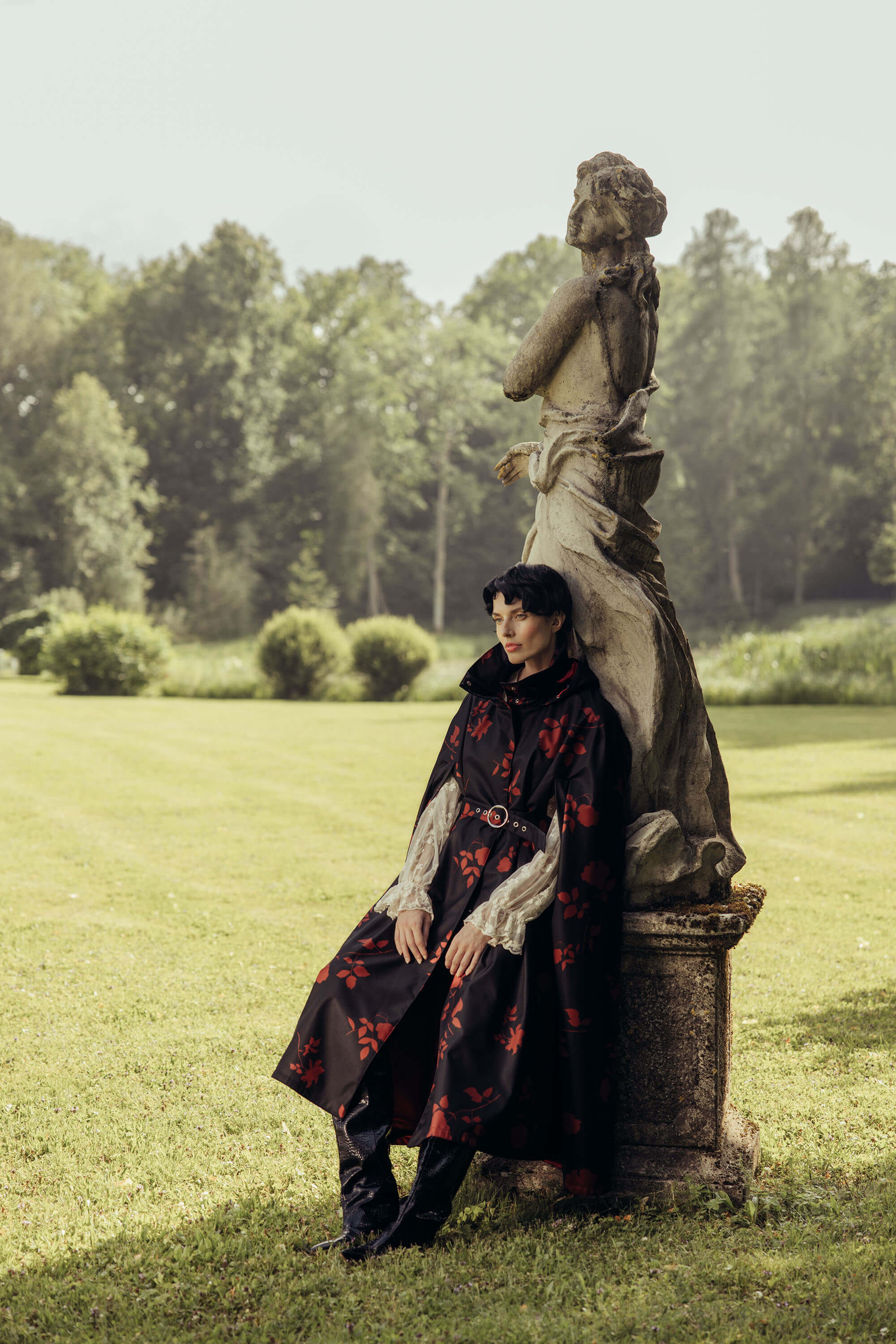 A person wearing a long black coat with red floral print by RainSisters styled with white blouse sits on a stone garden statue in a grassy park with trees in the background.