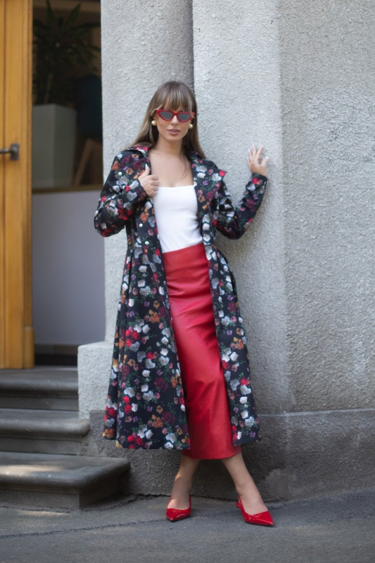 A model wearing a double-breasted trench coat with a floral pattern consisting of colorful vintage roses, paired with a white top and red skirt, standing outdoors.