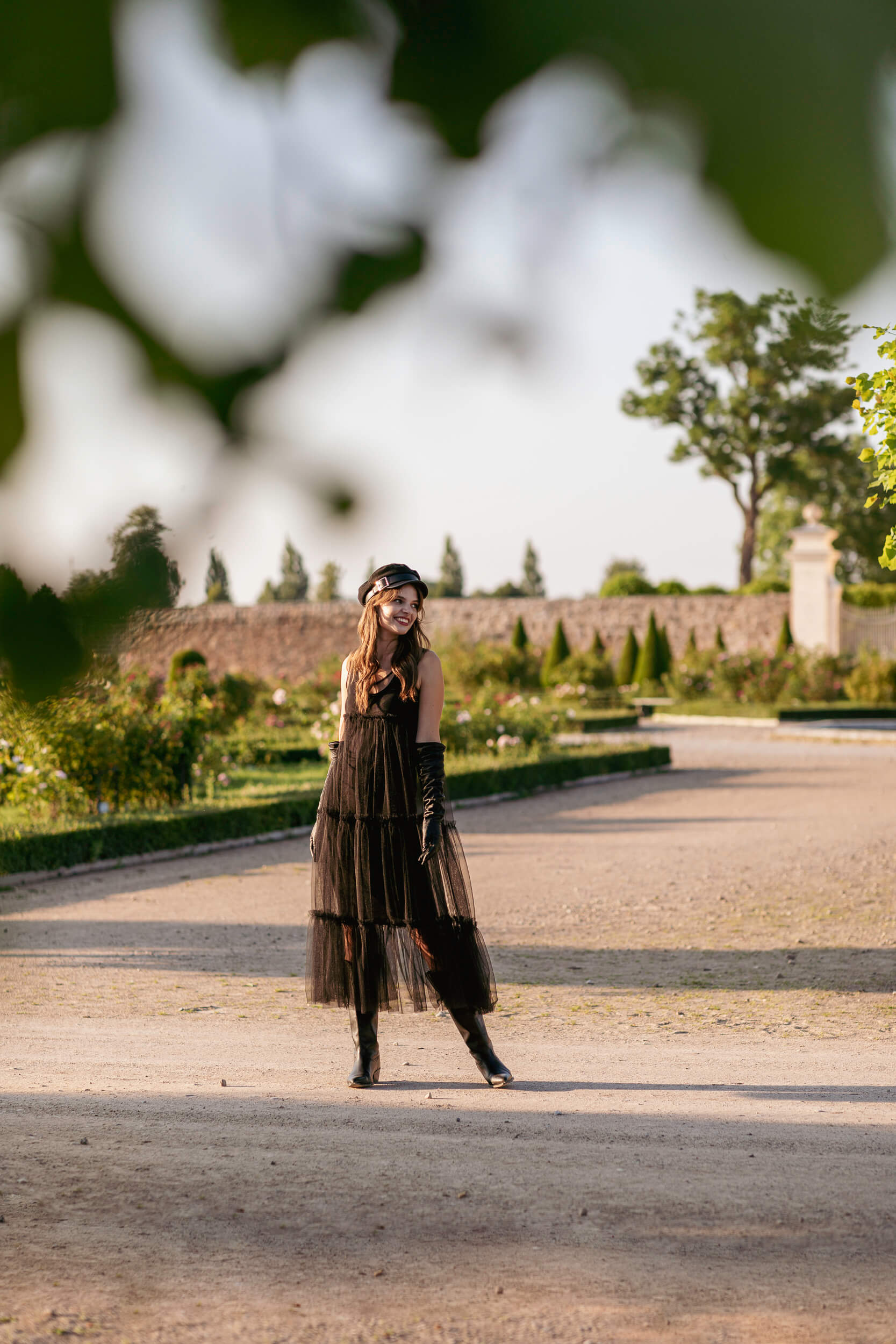 Woman in a long black dress standing on a path in a park with trees and a wall in the background.