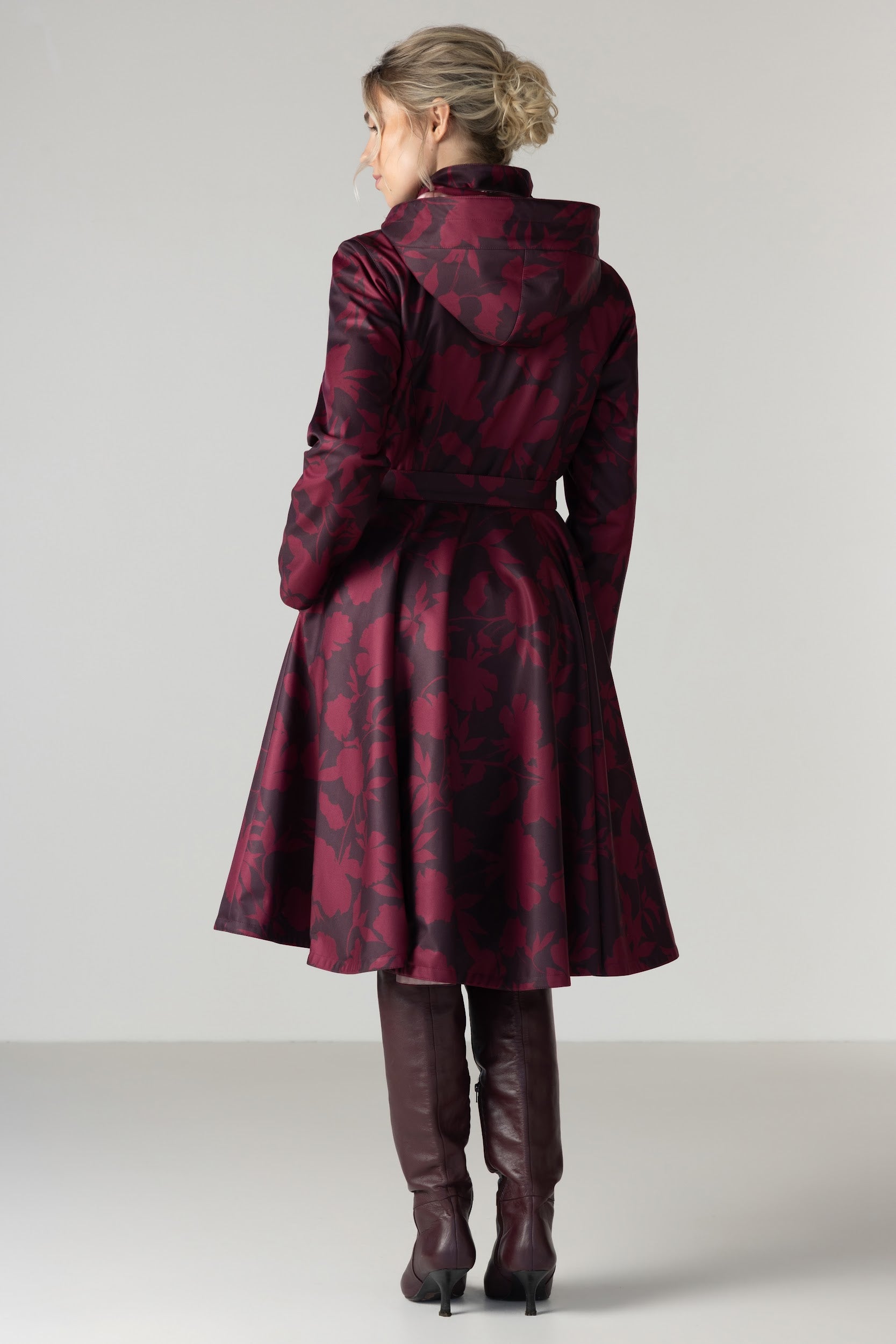 A woman viewed from the back wearing a dark red and black patterned hooded coat with a waist belt by RainSisters, paired with dark brown knee-high boots, against a light gray studio background.