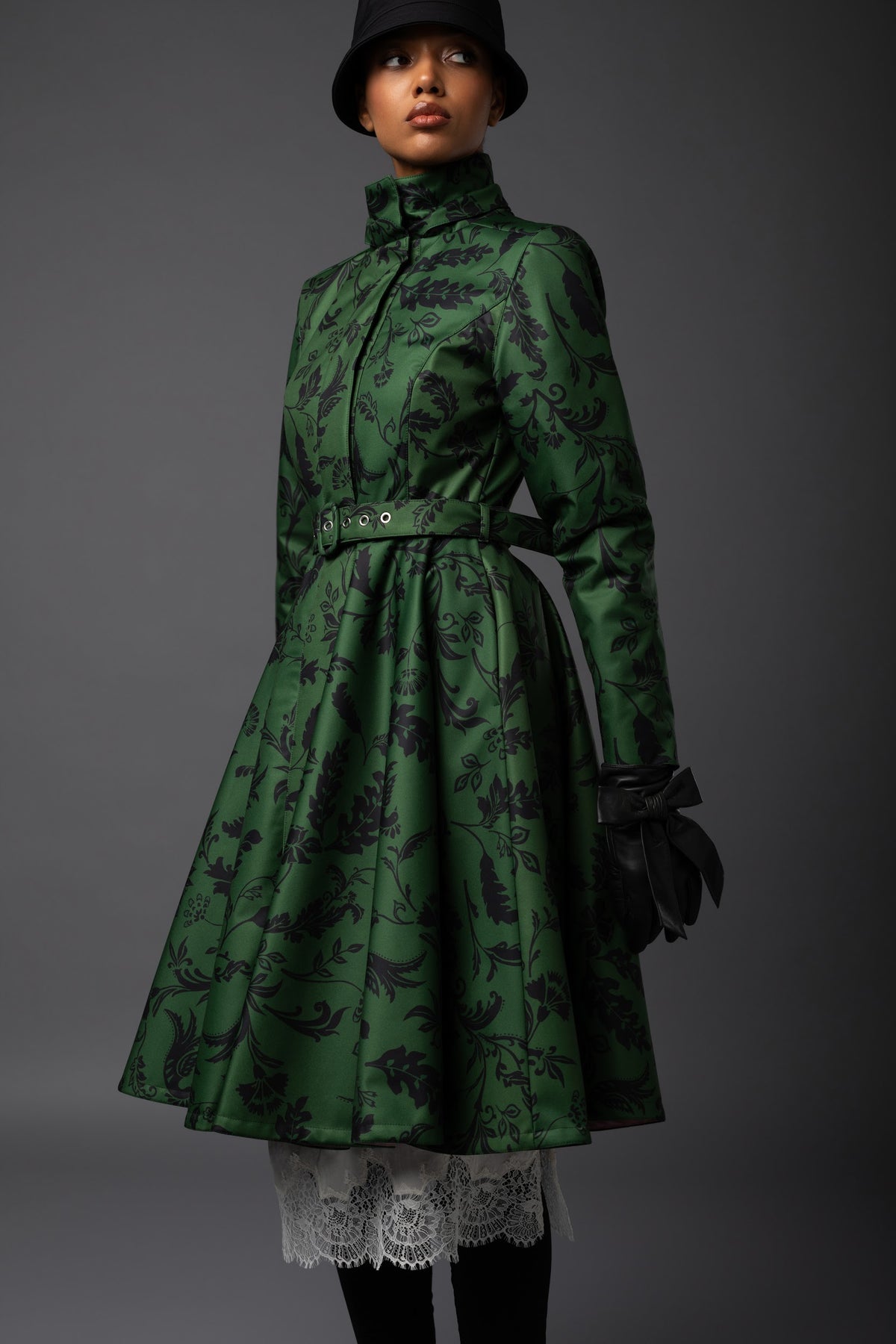 A model wears a dark green high-neck coat with a black floral pattern, styled with a matching belt, black gloves, a black hat, and a white lace hem, set against a dark gray studio background.