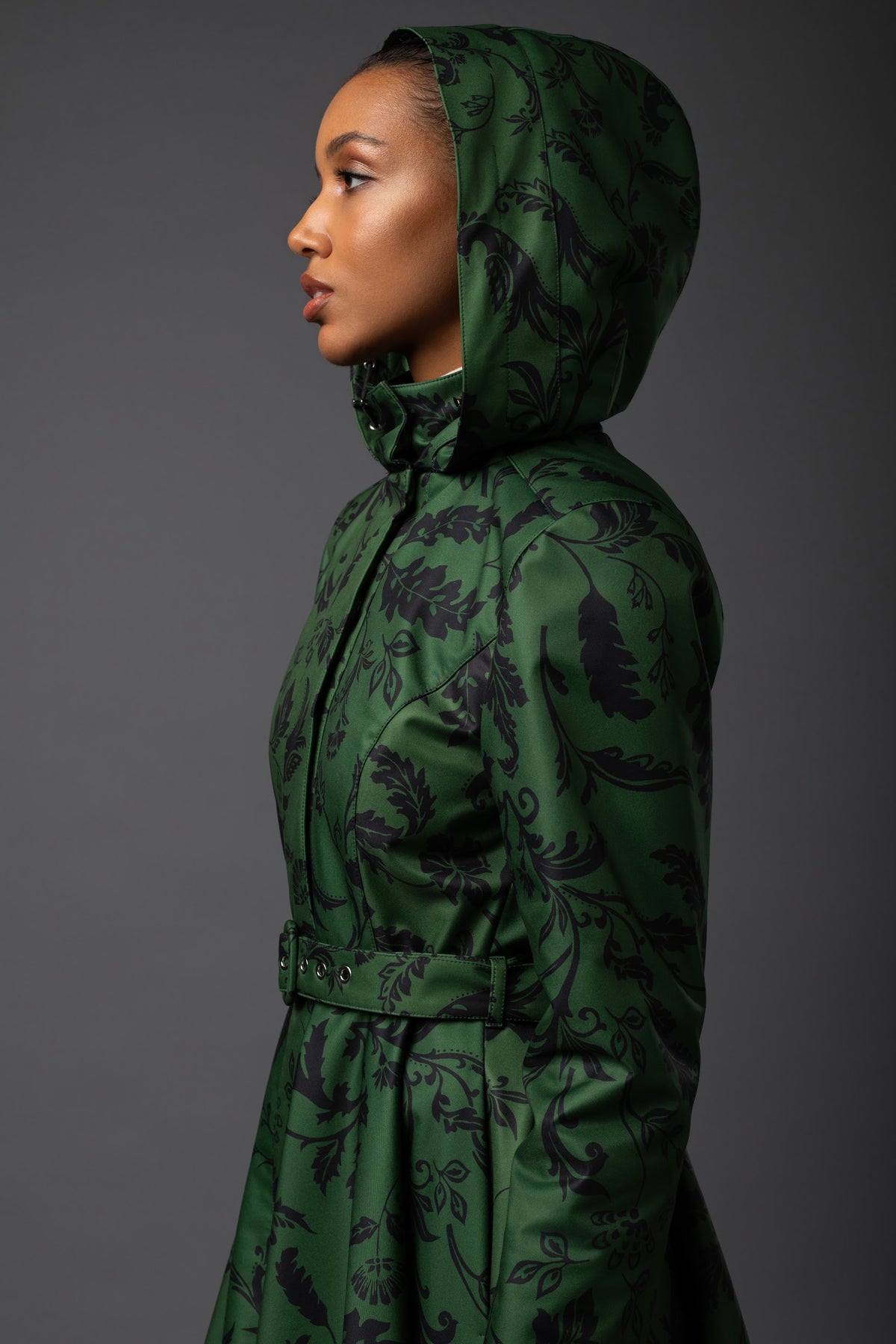 A model in profile wearing a Rainsisters winter dark green hooded raincoat with a black floral pattern, set against a solid dark gray studio background.
