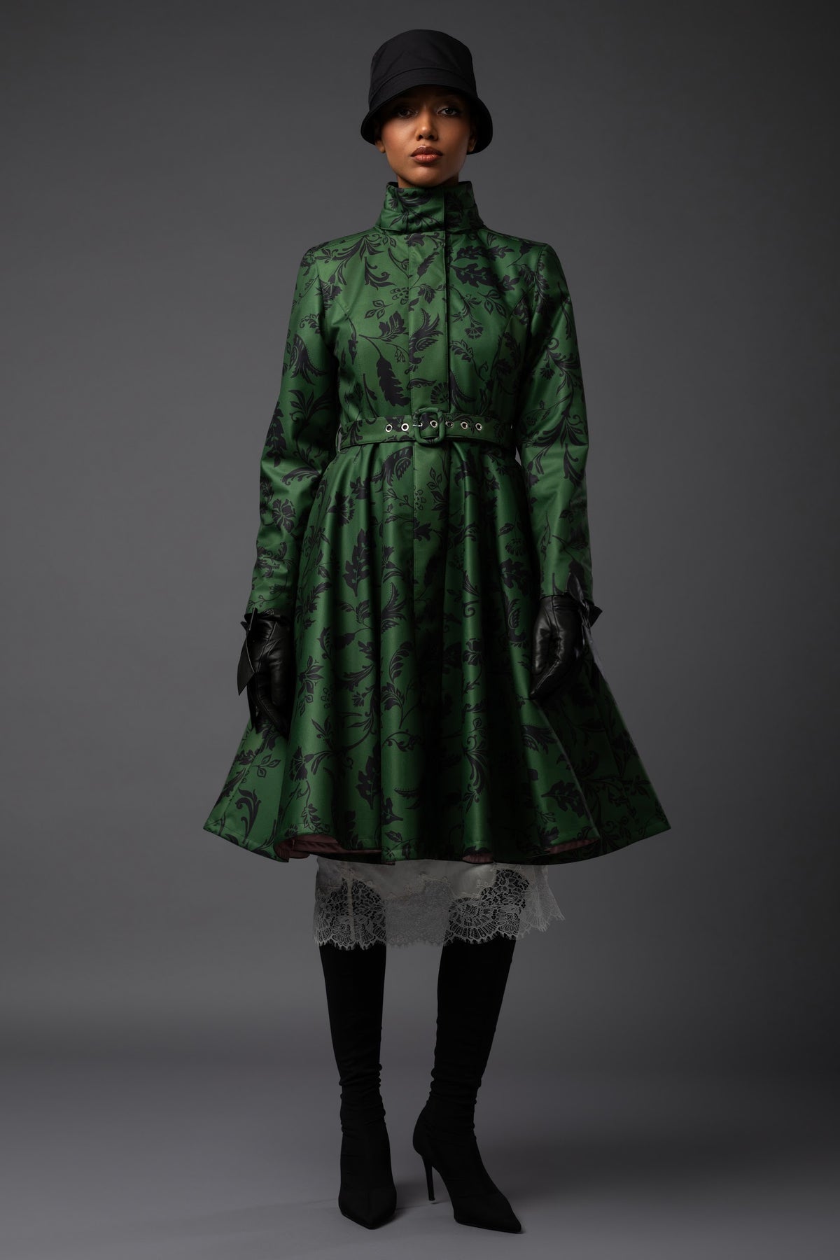 A model wearing a dark green patterned high-neck coat designed by Rainsisters with a matching buckle belt, black gloves, black knee-high boots, and a black hat, photographed against a dark gray studio background.