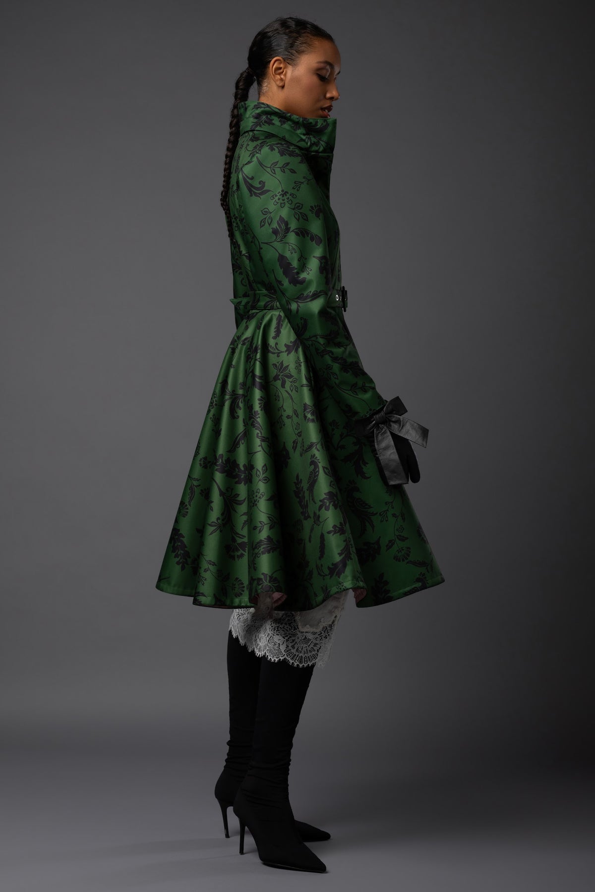 A model is shown from the side in a studio setting, wearing a green warm winter patterned high-neck waterproof coat with a waist belt, black knee-high boots.