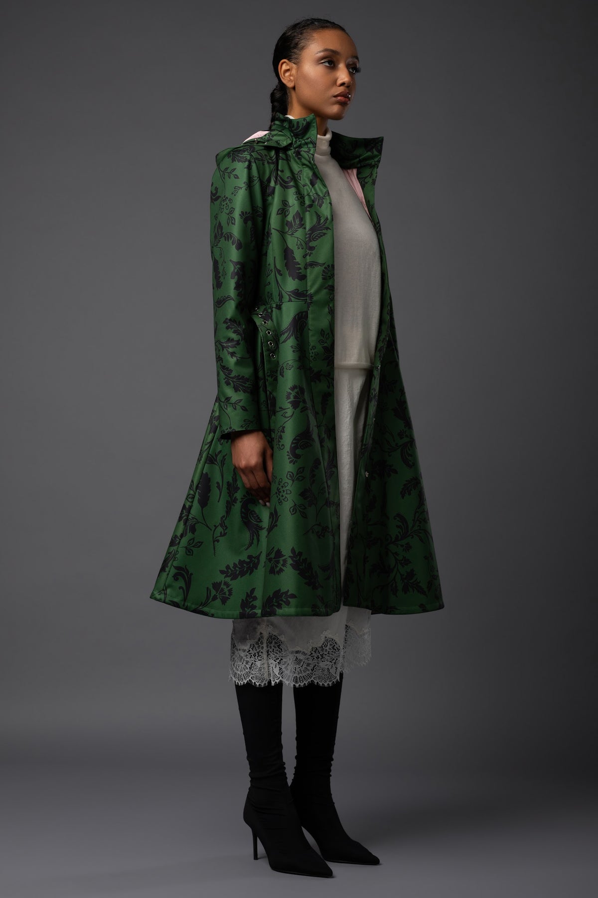 A model is shown from the side wearing Rainsisters long green waterproof coat with a black floral pattern, layered over a light dress, and black knee-high boots against a dark gray studio background.