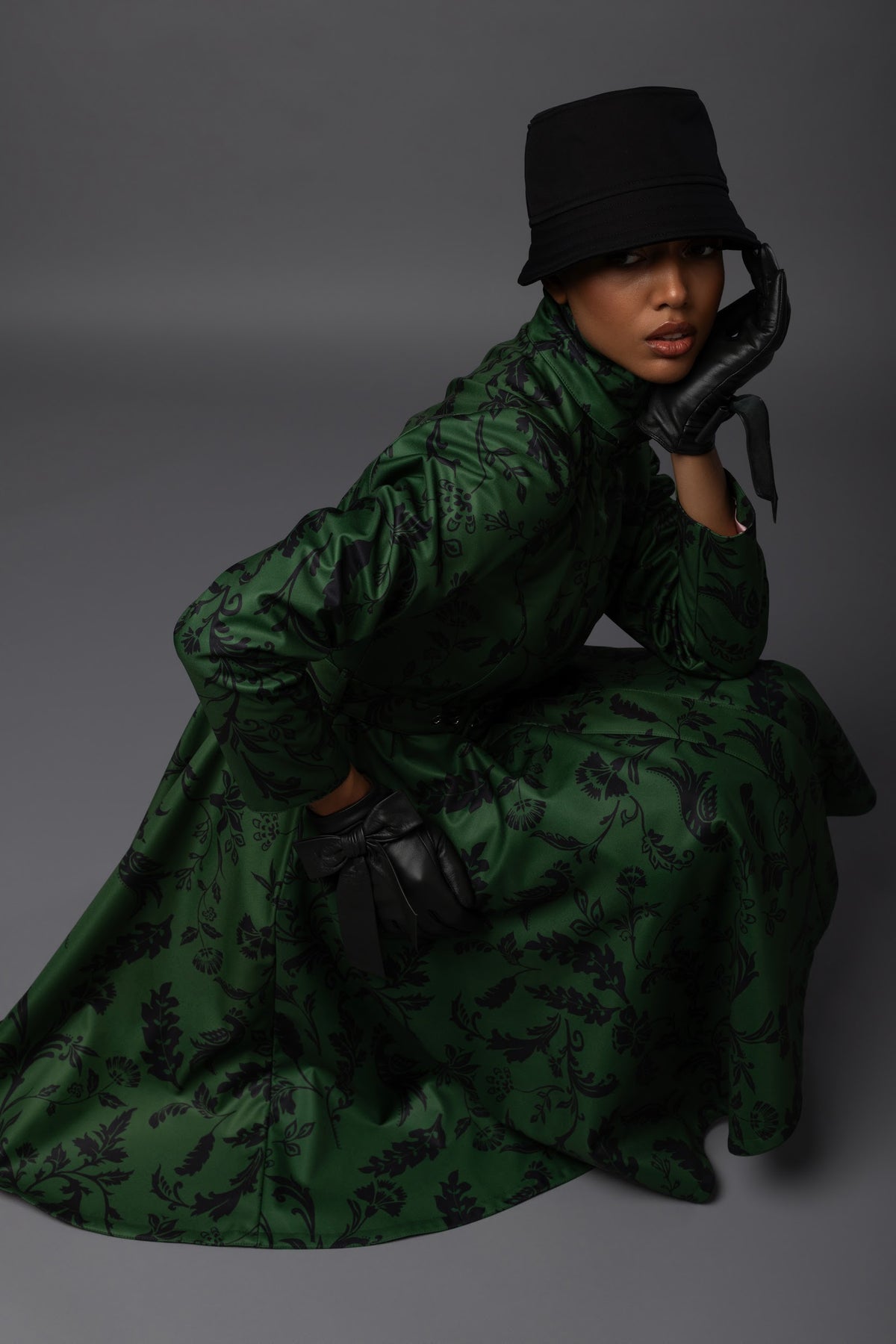 A model wearing a dark green long-sleeved waterproof winter coat with a black floral pattern, a black bucket hat, and black gloves, posed against a dark gray studio background.