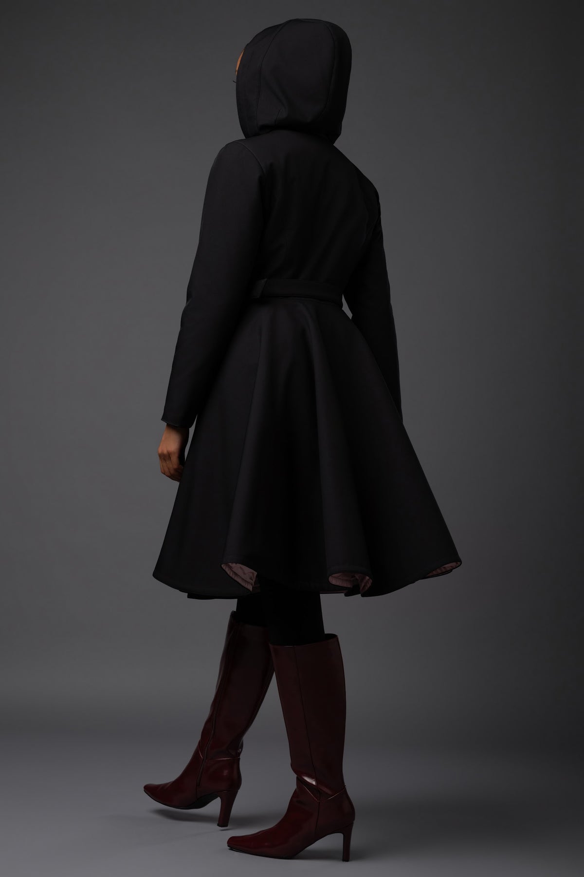 A model from behind wearing a black hooded winter longline Rainsisters coat with a flared skirt and a matching belt against a dark gray studio background.
