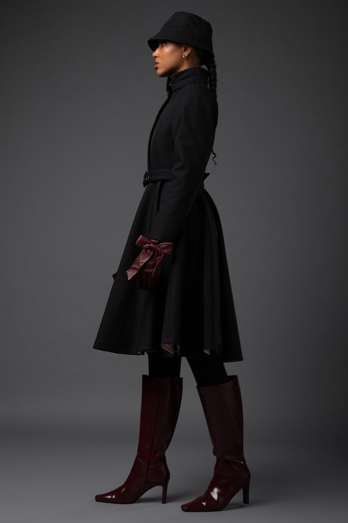 A model in profile wearing a Rainsisters black mid-length belted winter coat, a black hat, black leggings, and dark burgundy knee-high heeled boots against a dark gray studio background.