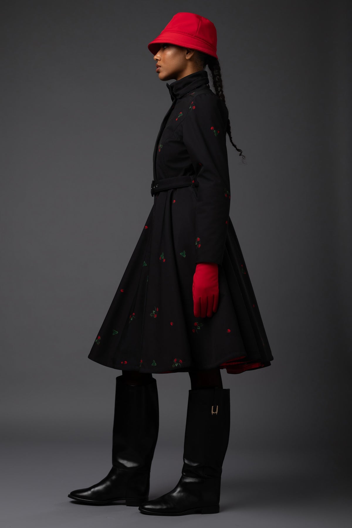 A model in profile wearing a black long waterproof winter coat with small red and green print details, a bright red bucket hat, a red glove, and black boots, set against a dark gray studio background.