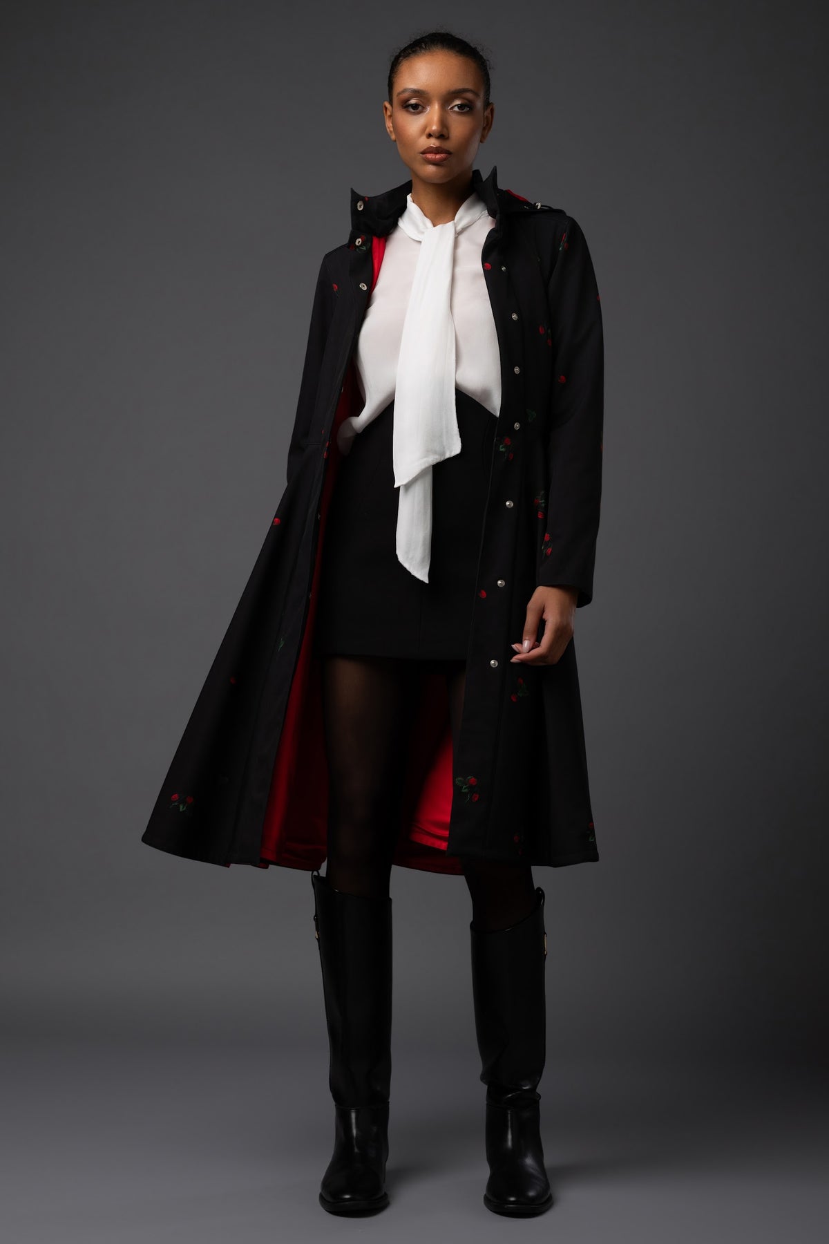 A model stands against a dark gray studio background wearing a long black winter overcoat with red lining and berry details, a white high-neck blouse, a black skirt, sheer black tights, and black knee-high boots.