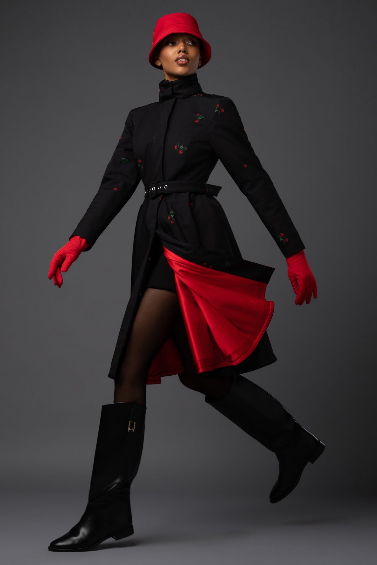 A model in a Rainsisters winter black coat with red berry details print, a red hat, red gloves, paired with black tights and black boots, captured in a dynamic pose against a dark gray studio background.