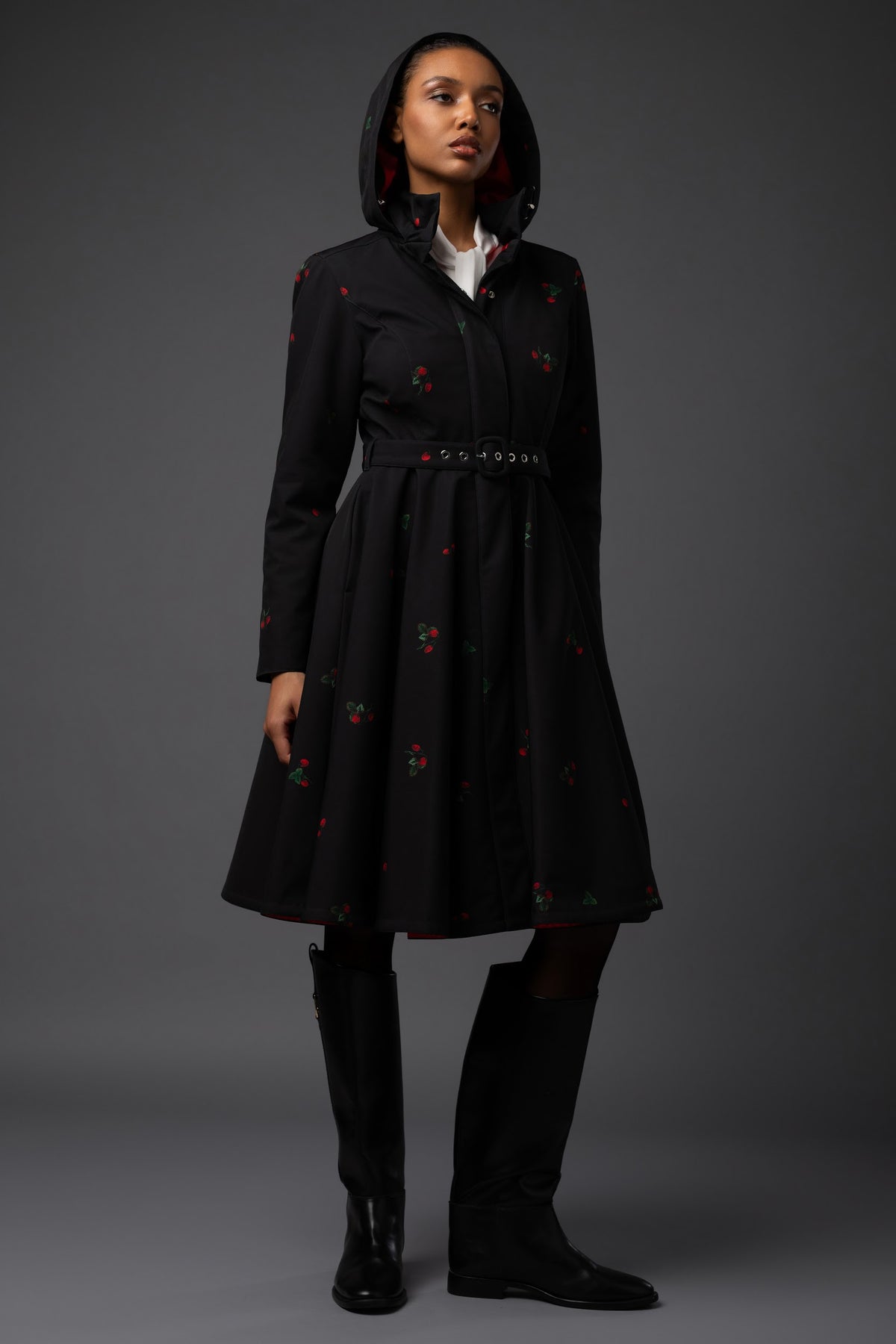 A model wears a black hooded longline warm winter coat with red and green floral details, styled with a white collared shirt, black knee-high boots, and black tights against a dark gray studio background.
