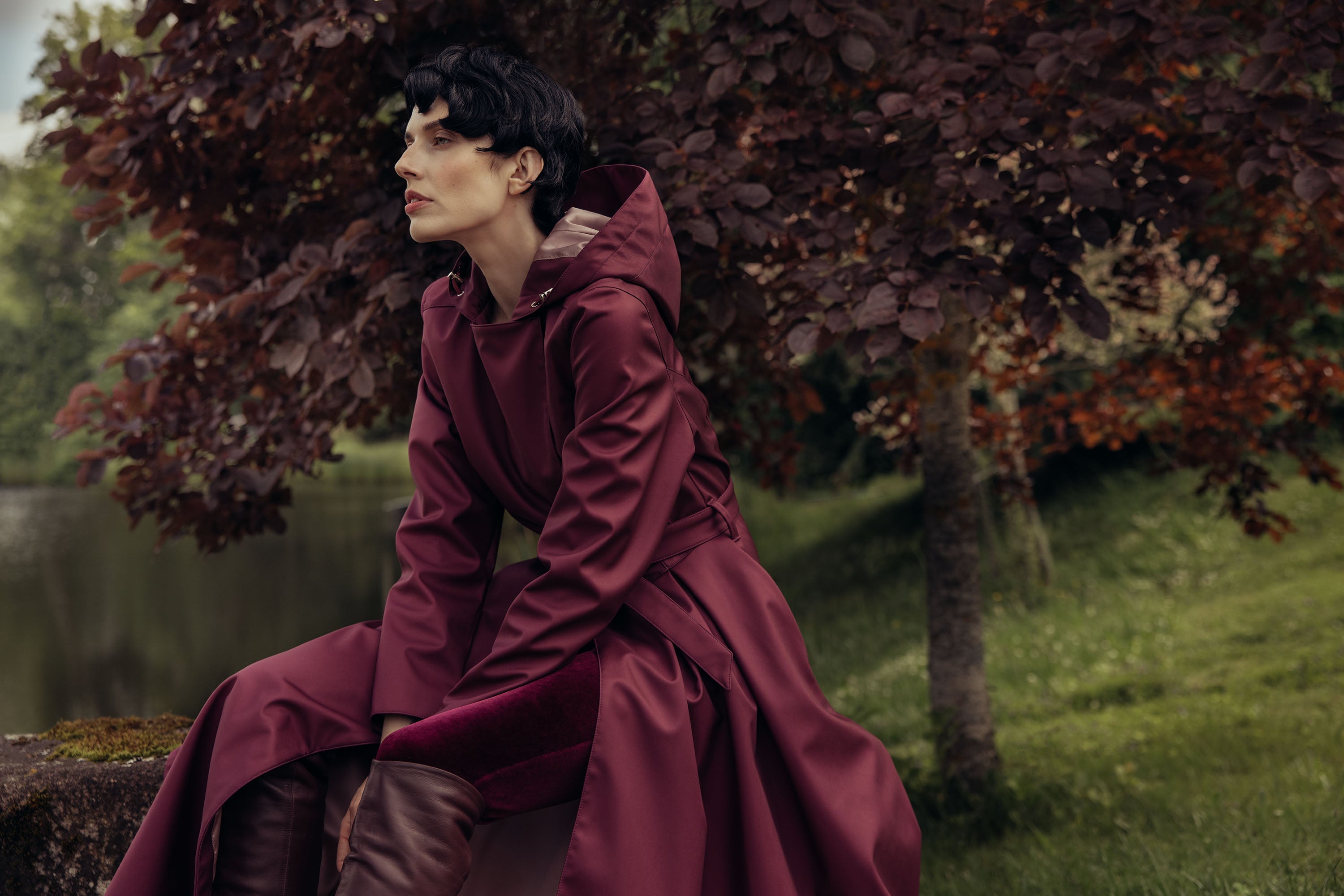 Model with short dark hair sitting on a rock wall wearing RainSisters waterproof coat Burgundy Night in Burgundy color