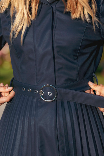 Blue pleated coat with a belt being adjusted against a blurred green background.