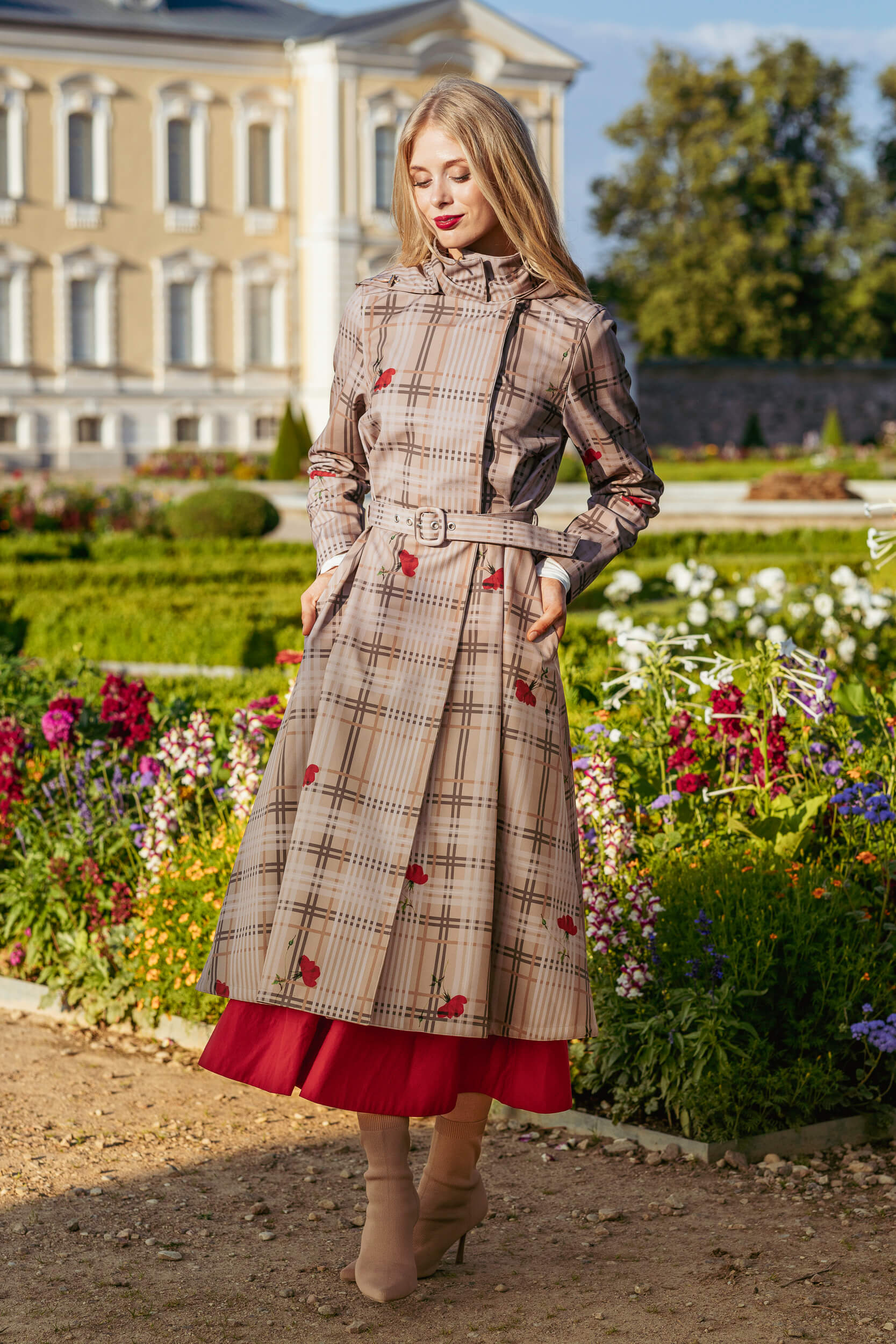 Woman in a plaid beige color coat with red accents standing in a garden with a classical building in the background