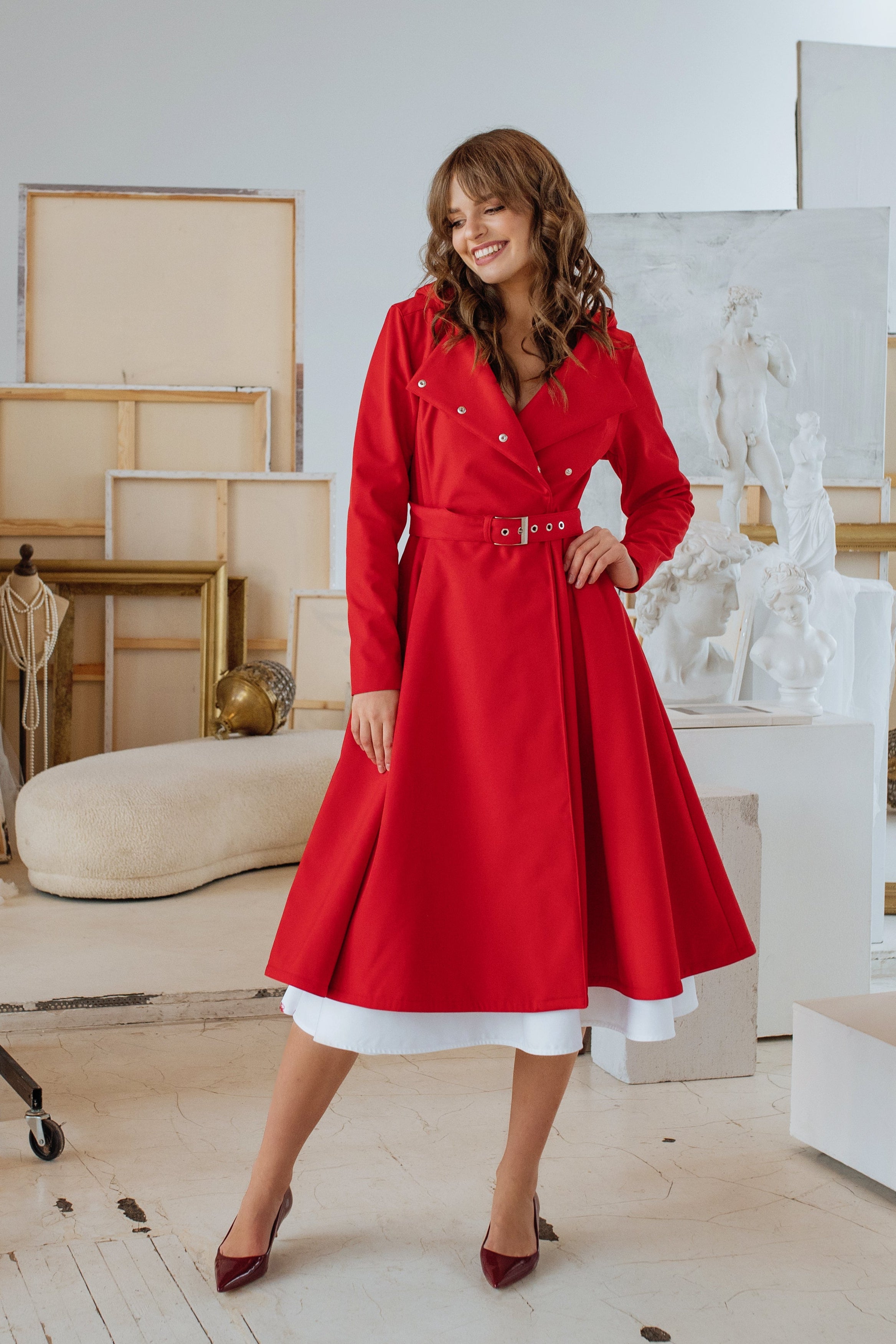 A model wearing a bright red double-breasted trench coat with a belt, featuring a midi length and snap button closure.