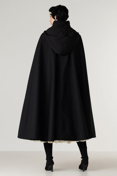 A model shown from the back wearing a black hooded cape coat by RainSisters, black tights, and black heels against a plain light gray studio background.