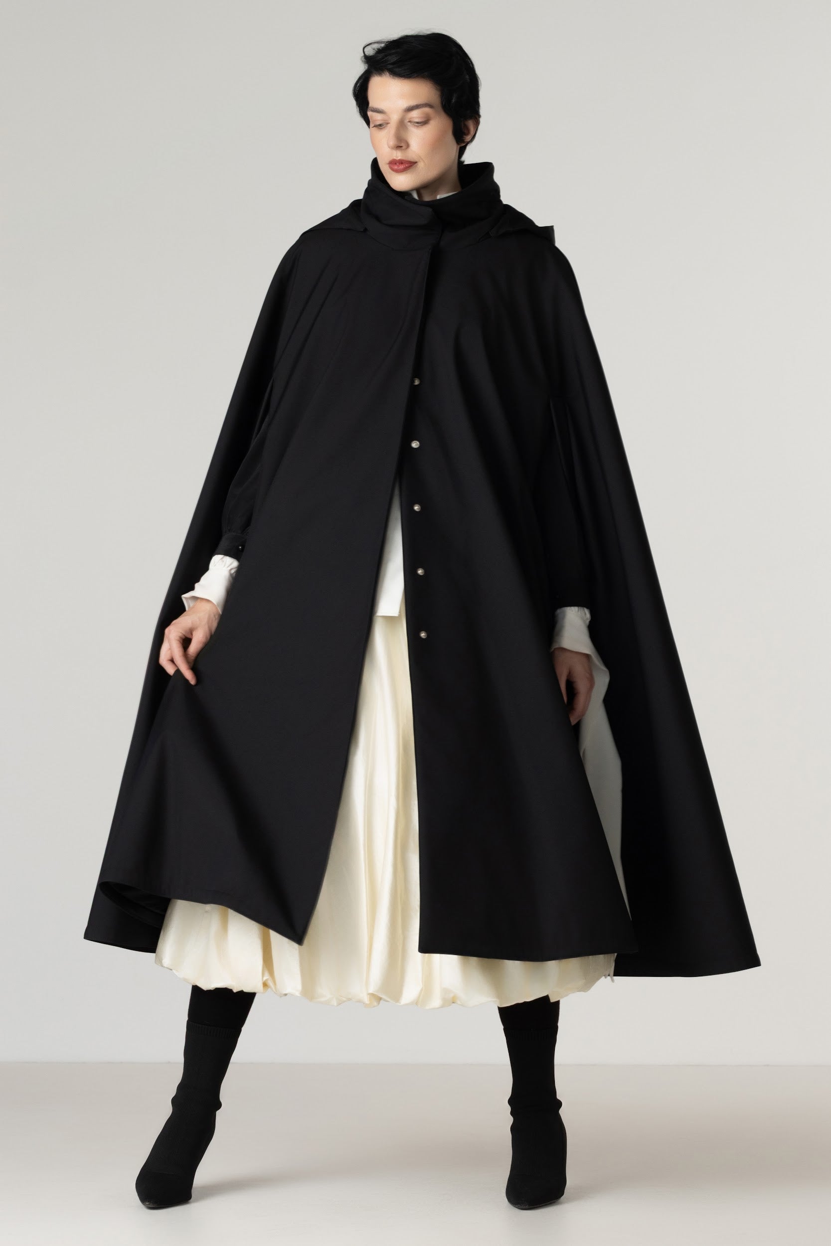 A model wearing a long black hooded cape-style coat by RainSisters over a cream pleated dress, black socks, and black boots, photographed against a plain light gray studio background.