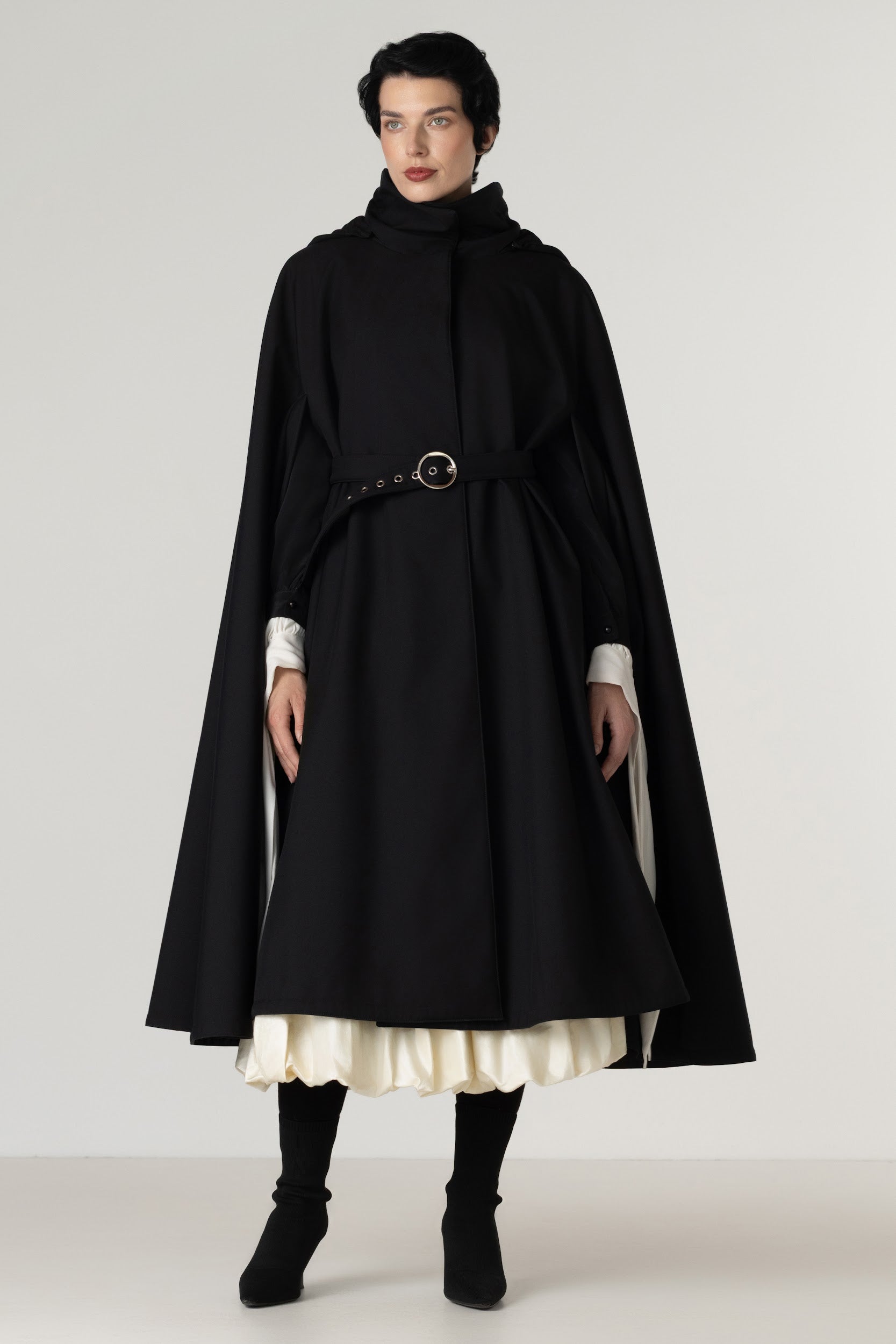 A model wearing a long black cape-style coat by RainSisters with a high mock neck, a matching waist belt with a round metal buckle, and layered white ruffled cuffs and hem, paired with black boots against a light gray studio background.