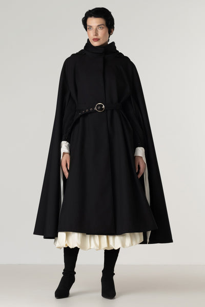 A model wearing a long black cape-style coat by RainSisters with a high mock neck, a matching waist belt with a round metal buckle, and layered white ruffled cuffs and hem, paired with black boots against a light gray studio background.