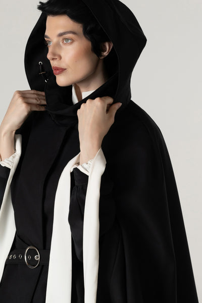 A model wearing a black hooded cape by RainSisters with a high collar against a light gray studio background.