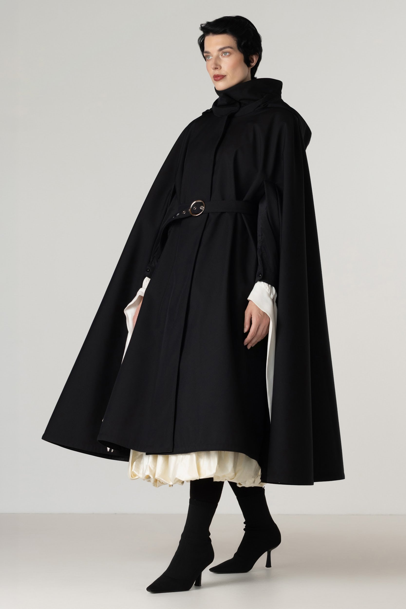 A model wearing a long black hooded cape coat by RainSisters with a matching waist belt, styled over a cream pleated dress and black knee-high boots, photographed against a neutral light gray studio background.