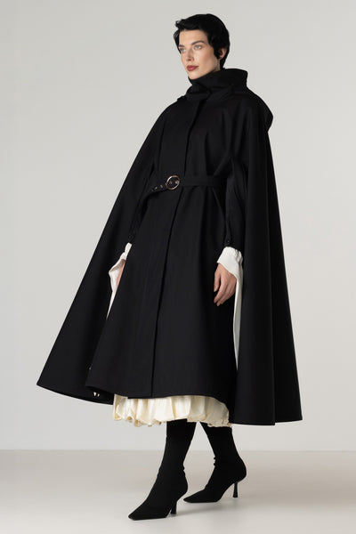 A model wearing a long black hooded cape coat by RainSisters with a matching waist belt, styled over a cream pleated dress and black knee-high boots, photographed against a neutral light gray studio background.