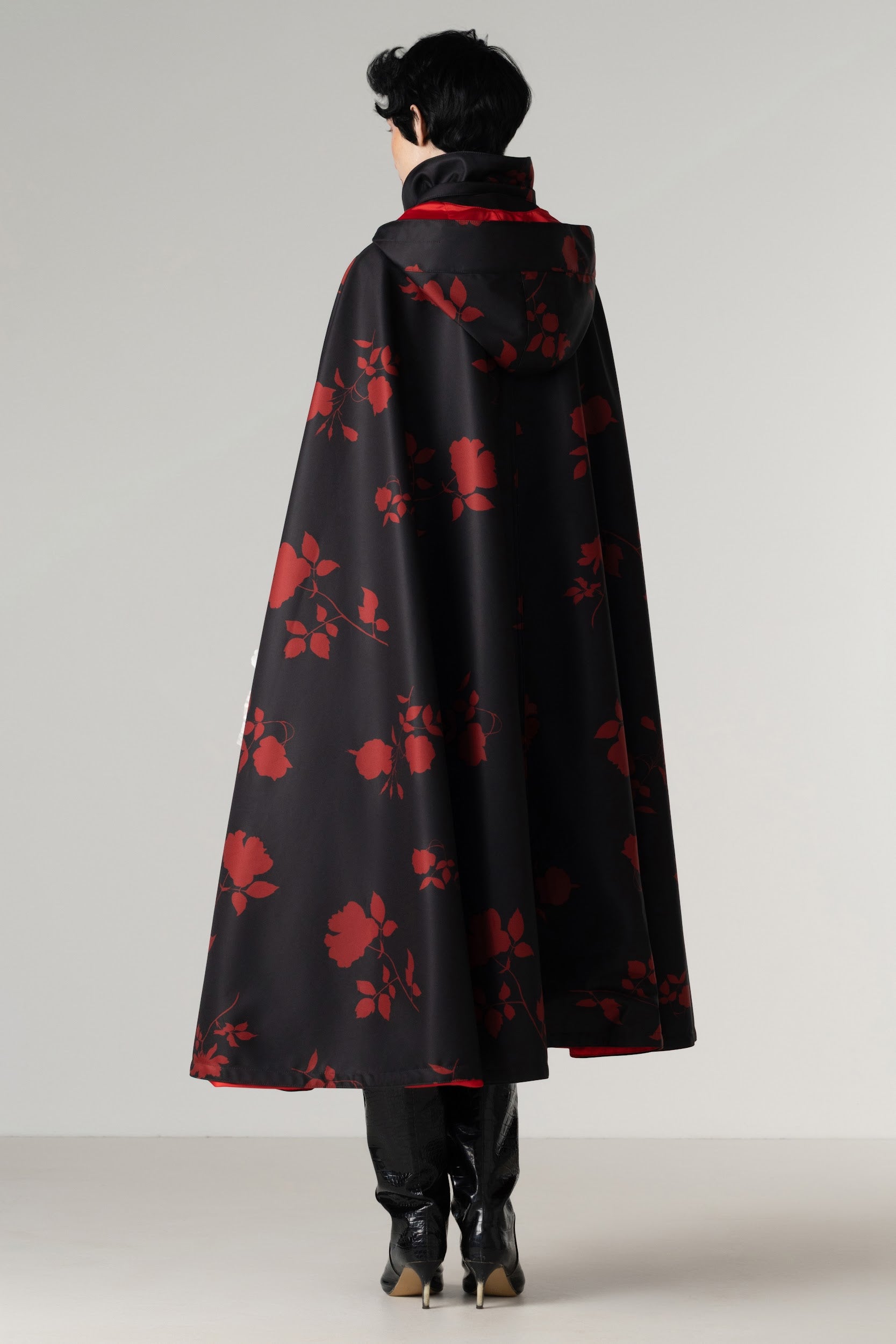 A model viewed from the back wearing a black hooded cape with a red floral pattern and a red inner lining, paired with black boots, against a light gray studio background.