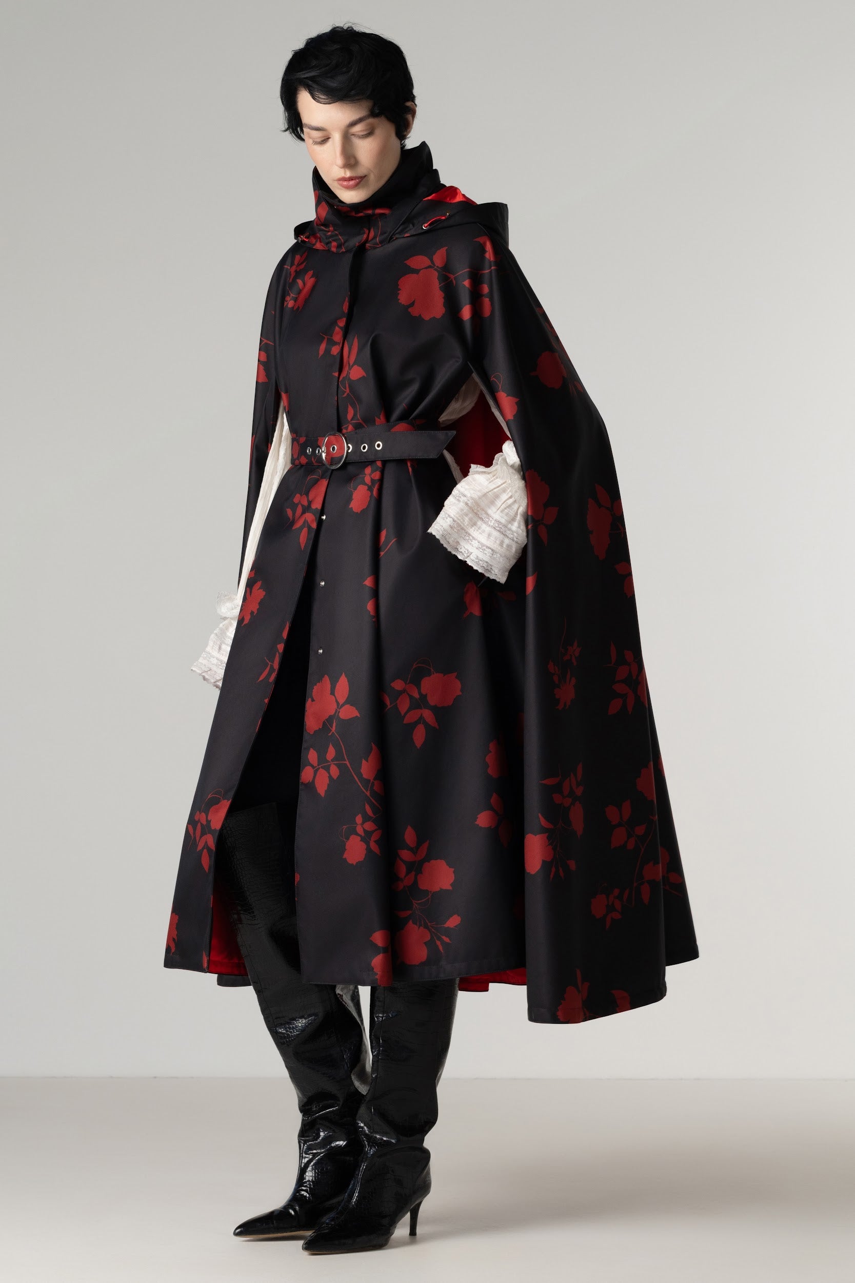 A model wearing a long black cape-style coat by RainSisters with a red floral pattern, a matching belt, black trousers, and black pointed-toe boots against a light gray studio background.