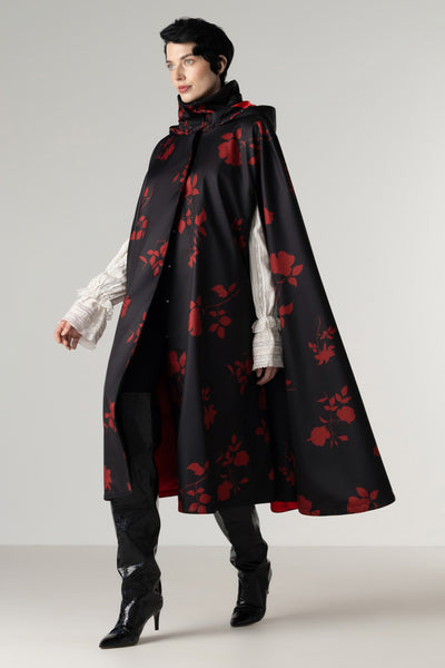 A model wearing a long black cape with a red rose pattern by RainSisters, styled over a white ruffled shirt, black pants, and black high-heeled boots, set against a plain light gray studio background.