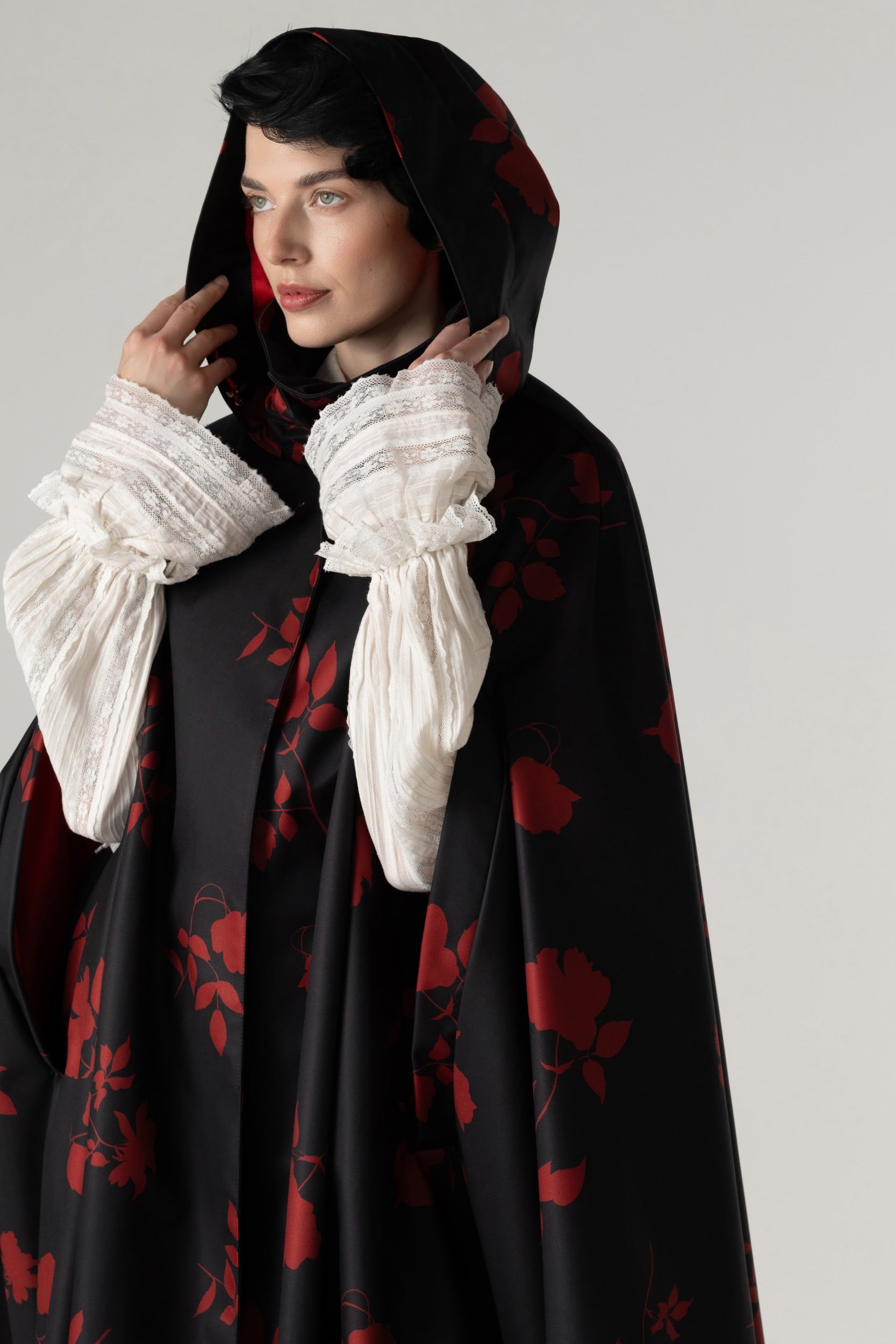 A model wearing a black hooded cape with a red floral pattern by RainSisters, styled with dramatic white ruffled sleeves, against a neutral light gray background.