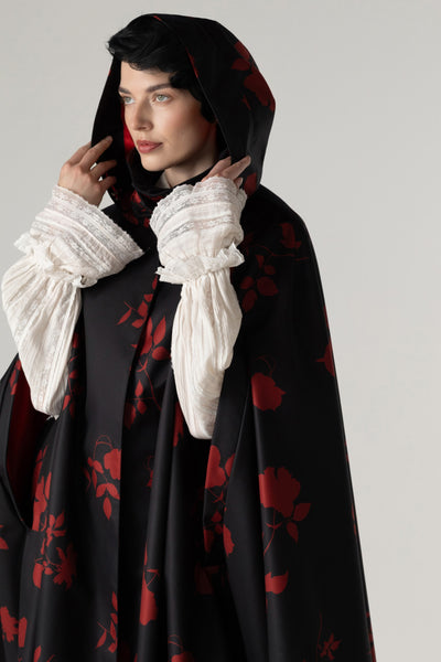 A model wearing a black hooded cape with a red floral pattern by RainSisters, styled with dramatic white ruffled sleeves, against a neutral light gray background.