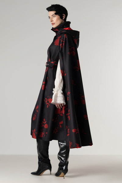 A model is shown from the side wearing a long black hooded cape with a red floral pattern by RainSisters, styled over a white ruffled long-sleeve blouse, black pants, and black high-heeled boots against a light gray studio background.