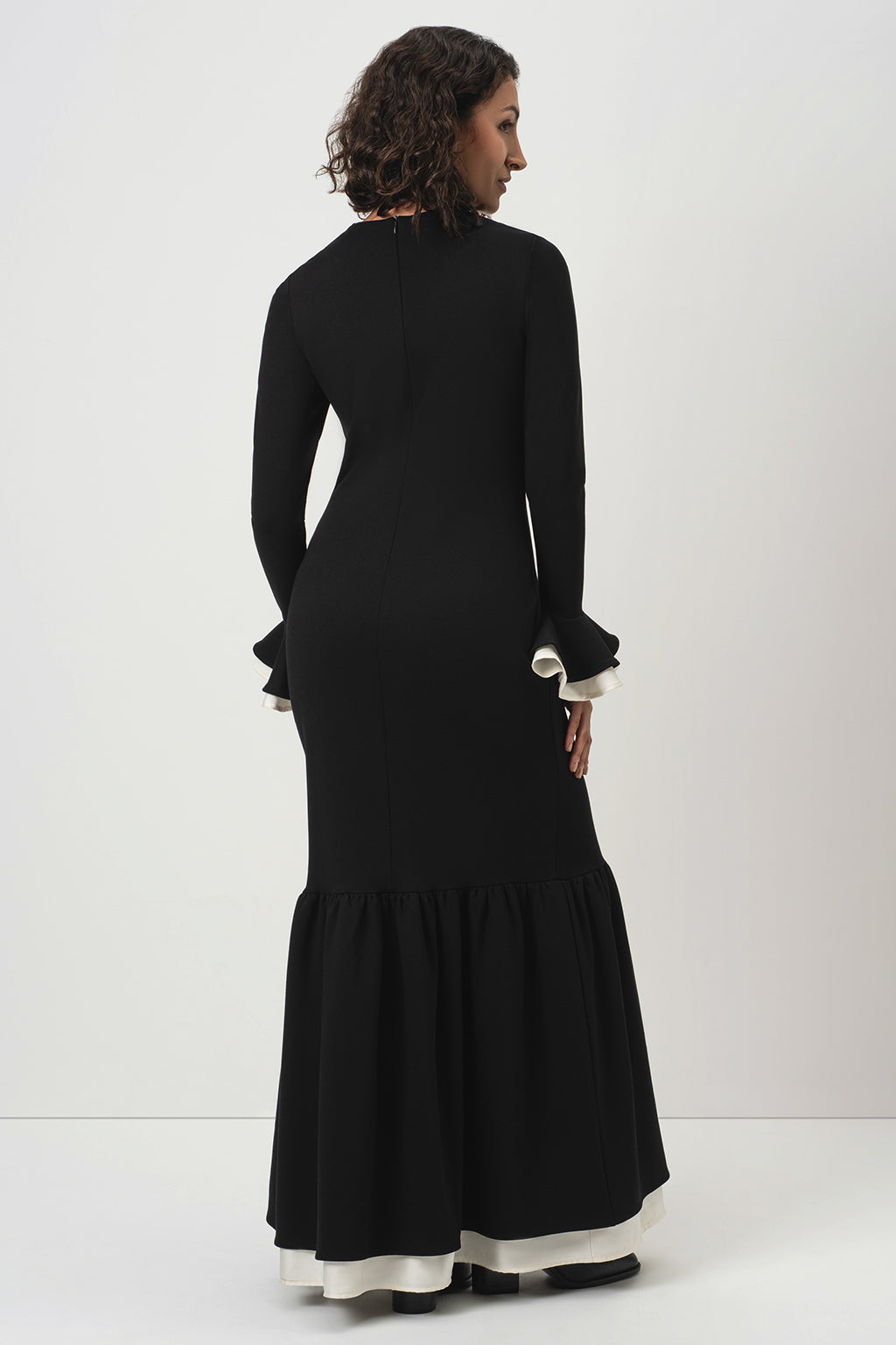 A woman wearing RainSisters long black dress Quiet Elegance with long sleeves, ruffled cuffs, and a flared skirt, shown from the back against a plain light gray studio background.