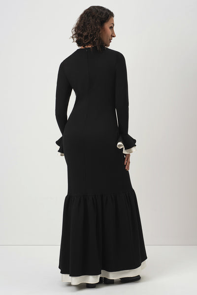 A woman wearing RainSisters long black dress Quiet Elegance with long sleeves, ruffled cuffs, and a flared skirt, shown from the back against a plain light gray studio background.