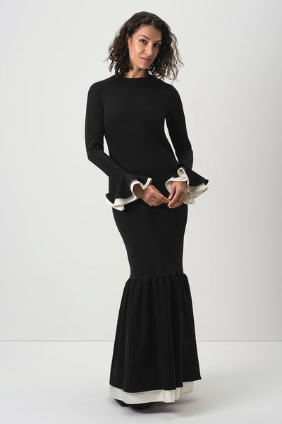 A woman modeling RainSisters black long-sleeve mermaid-style dress Quiet Elegance with contrasting white ruffle trim at the cuffs and hem, photographed against a plain light gray studio background.