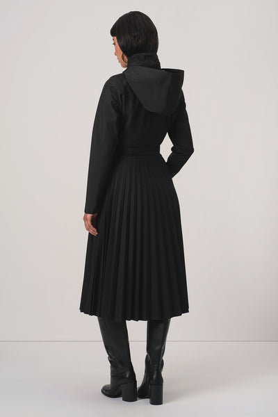 A model viewed from the back wearing RainSisters black hooded long-sleeve coat Black Sway with a pleated skirt and black knee-high boots, set against a plain light gray studio background.