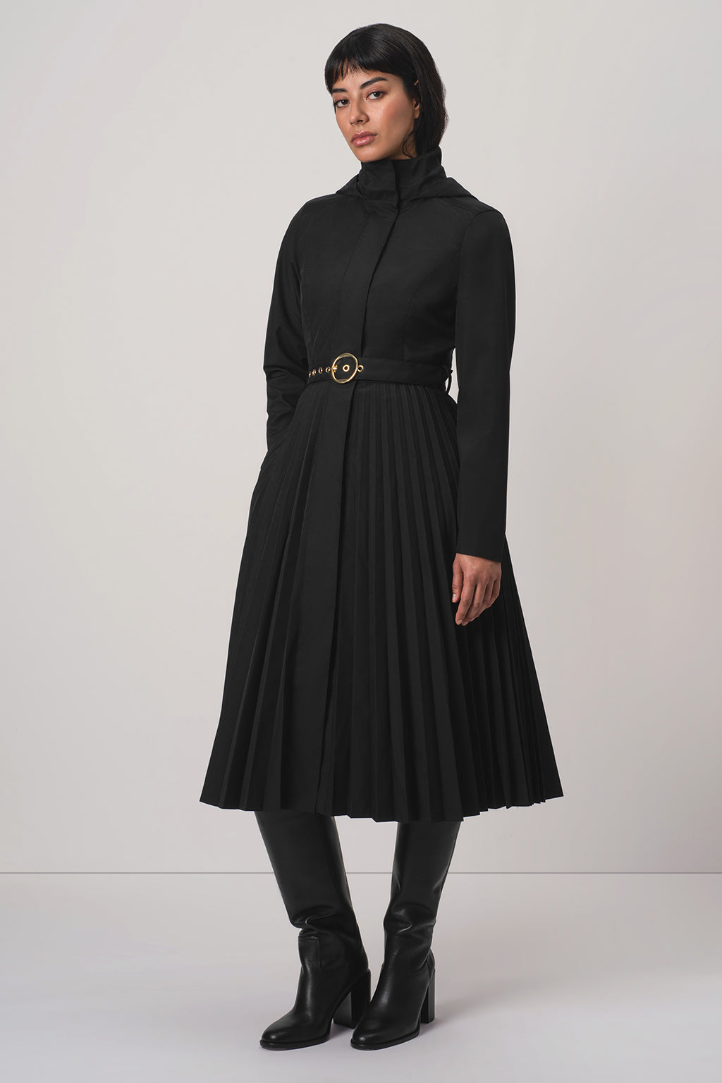 A model wearing RainSisters black long-sleeve midi coat Black Sway with a pleated skirt and a gold-tone belt, paired with black knee-high boots, against a light gray studio background.