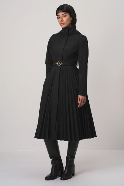 A model wearing RainSisters black long-sleeve midi coat Black Sway with a pleated skirt and a gold-tone belt, paired with black knee-high boots, against a light gray studio background.