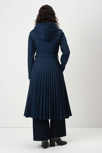 A model viewed from the back wearing RainSisters dark navy hooded coat Blue Serenade with a pleated skirt, wide-leg trousers, and black boots against a plain white studio background.