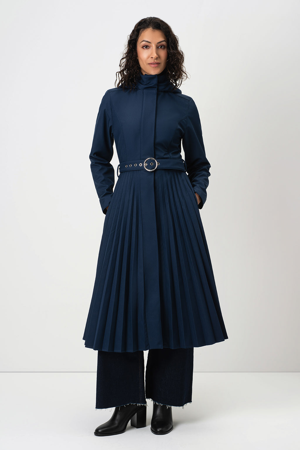 A woman modeling RainSisters dark navy blue long-sleeve trench coat Blue Serenade with a high mock neck, a detachable hood, a thin belt with a round metal buckle, wide-leg trousers, and black ankle boots, set against a plain light gray studio background.